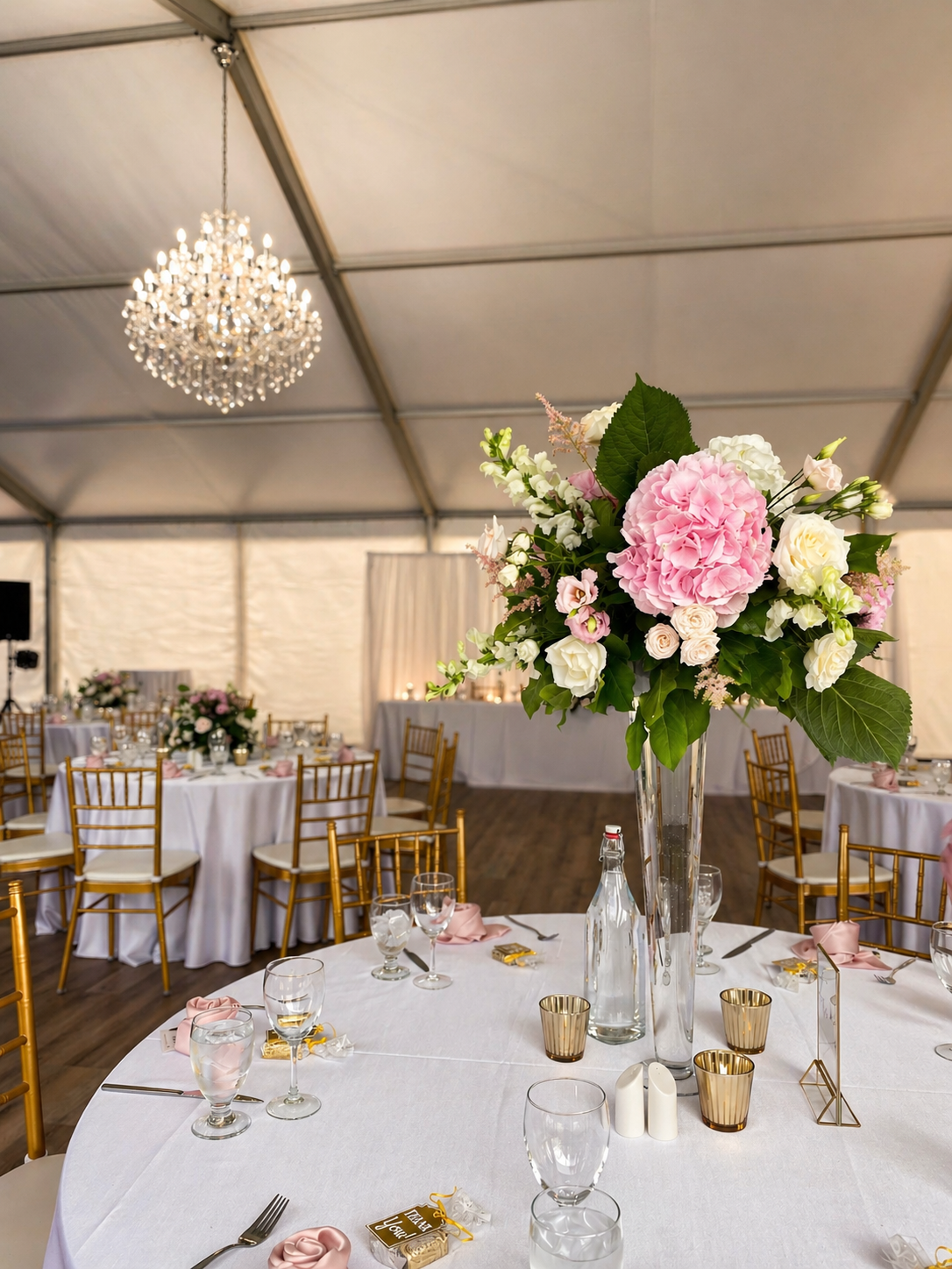 Lavish floral centerpiece featuring premium blooms on an impeccably styled reception table inside the Waterfront Pavilion tent, illuminated by an elegant crystal chandelier.