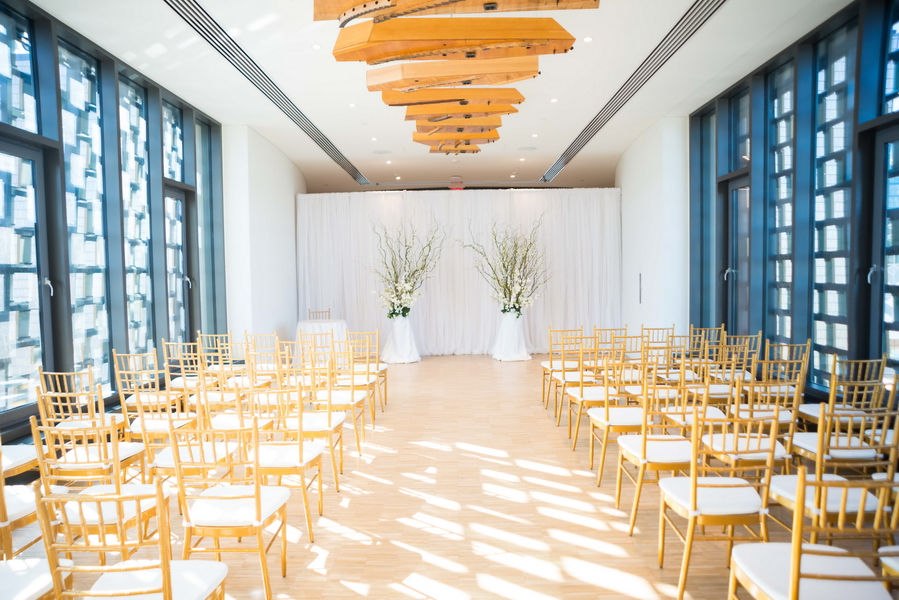 Beautifully styled wedding ceremony aisle lined with sophisticated seating in a bright, sunlit event space, demonstrating exceptional scenic production and atmospheric design.